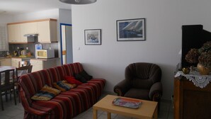 A living room with a sofa, armchair, and a small table. There is a kitchen area with a microwave and cabinets. Two framed pictures are on the wall. - Detached house, in an enclosed garden, quiet location, near forest and sea (Saint-Vincent-sur-Jard)