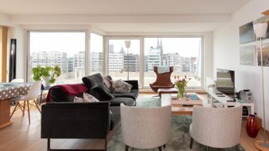 A modern living room with a sofa, two armchairs, a coffee table, and a dining area with a table and chairs. Large windows offer a view of the cityscape. - Sunny Apartment With Large Terrace (Ostend)