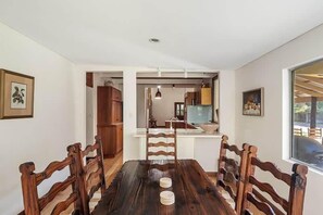 A dining area with a wooden table and chairs, a kitchen in the background, and a window with a view. - Orchard Homestead—A True Rural Wellness Escape (Yallingup Siding)
