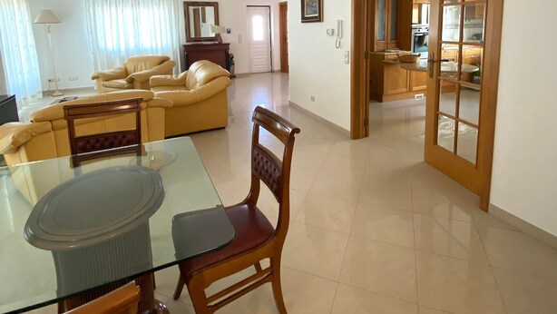 A spacious living area with a glass dining table, wooden chairs, a sofa, and a kitchen visible through an open door. - Stunning Town Villa close to beach and Golf courses (Caldas da Rainha)