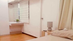 A bedroom with a bed, a bedside table with a lamp, a small window with blinds, and a white ceiling. - 2bedroom condo in enjoyable Quezon City with WiFi, AC (Quezon City)