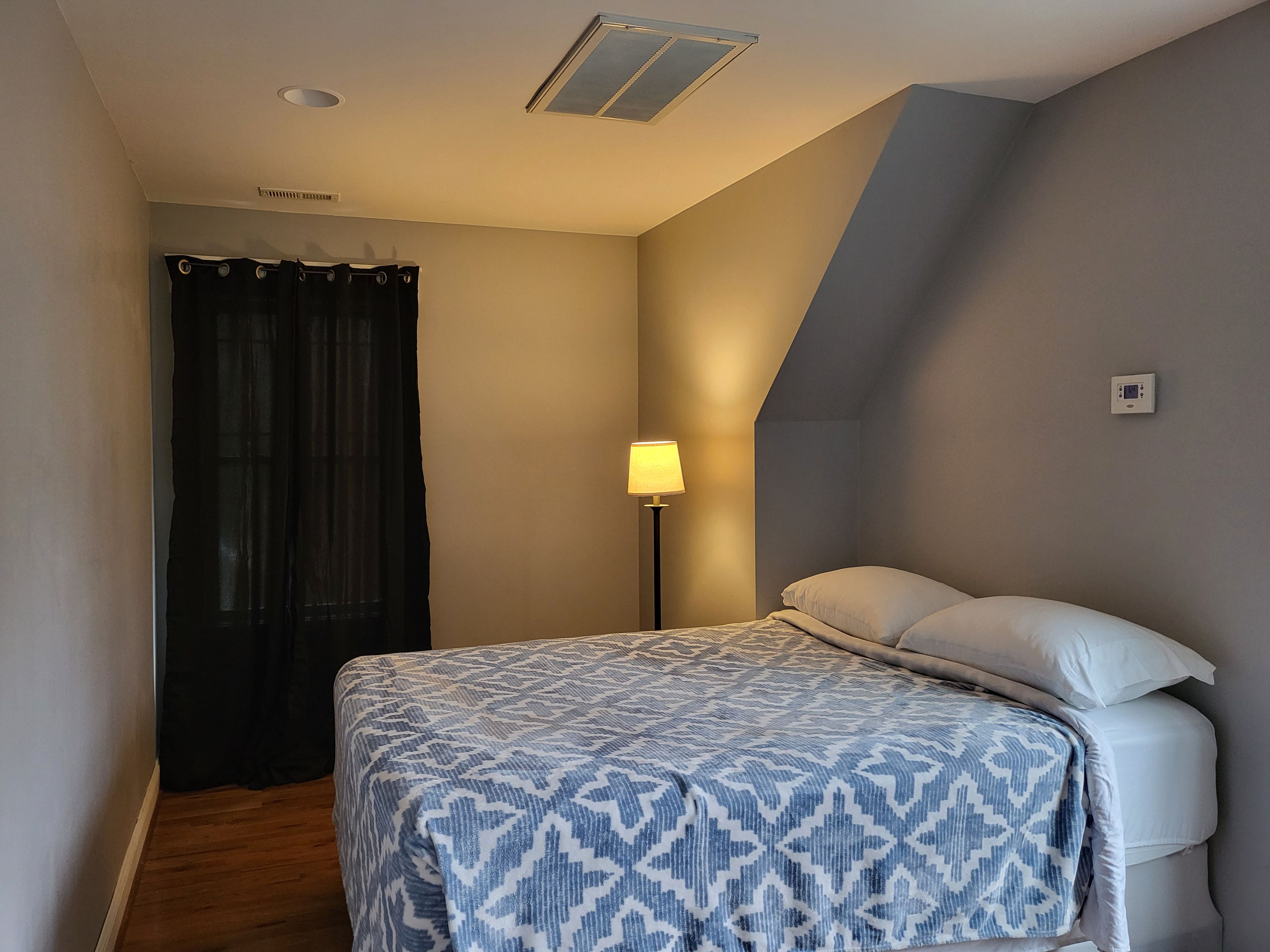 Une chambre à coucher avec un lit, une table de chevet, une lampe, une fenêtre avec des rideaux et un thermostat fixé au mur.