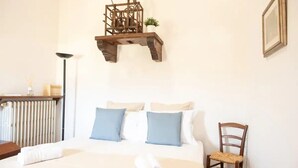 Una cama bien hecha con almohadas azules y blancas, una silla de madera, un radiador, una lámpara de pie y un estante en la pared con una planta.