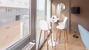 A modern hotel room with large windows, a round mirror, a small round table with chairs, and a blue sofa. - Apartment With Side sea View (Ostend)