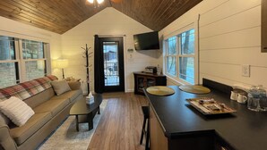 Uma sala de estar acolhedora com teto de madeira, um sofá, uma mesa de centro e uma área de cozinha com bancada e televisão.