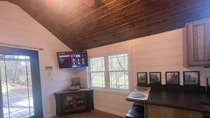 Interior de cabana com teto de madeira, TV na parede, área de bar com banquetas e quadros decorativos.