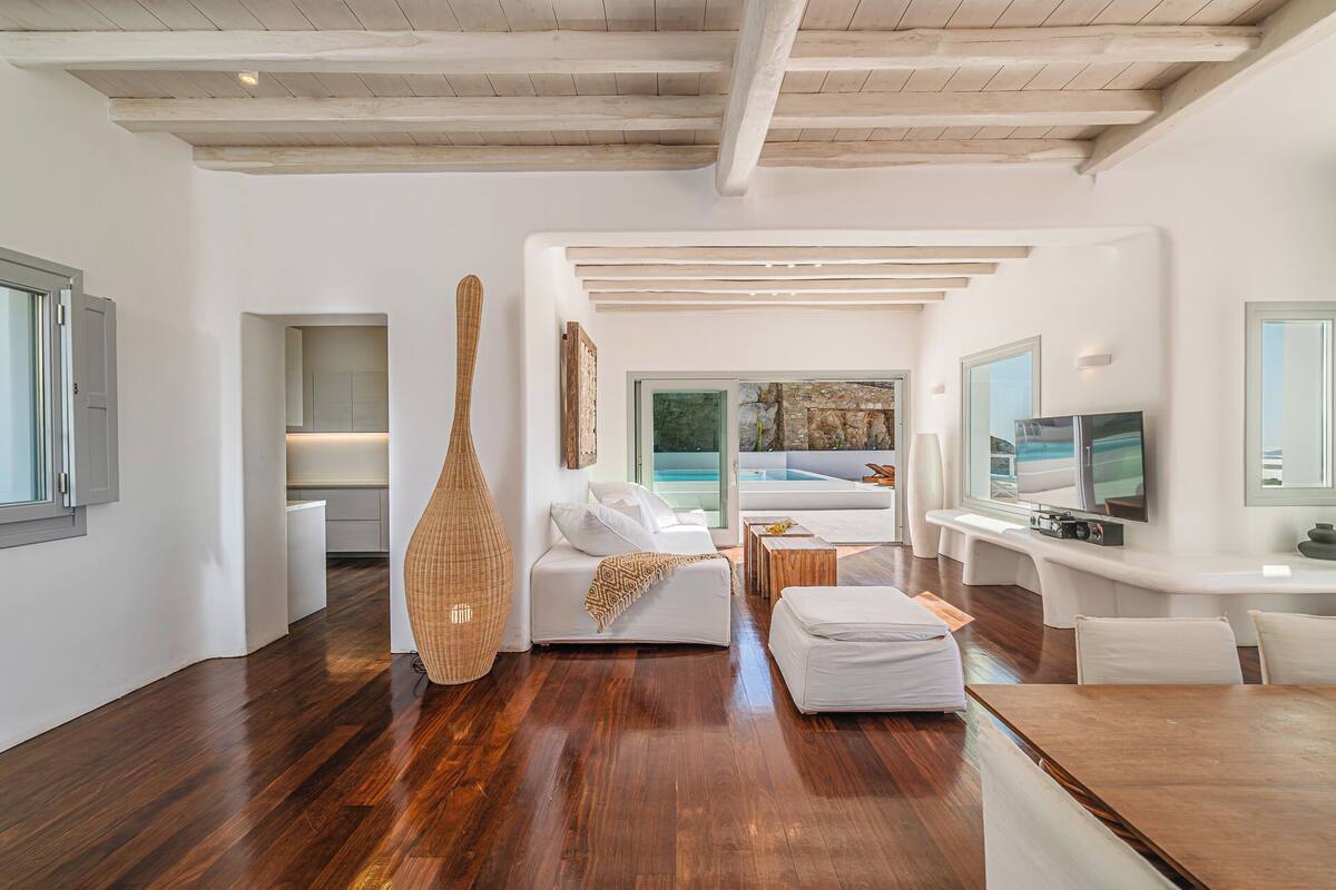 Un amplio salón con techo de madera, un ventanal con vistas a una piscina y un televisor sobre un mueble blanco.