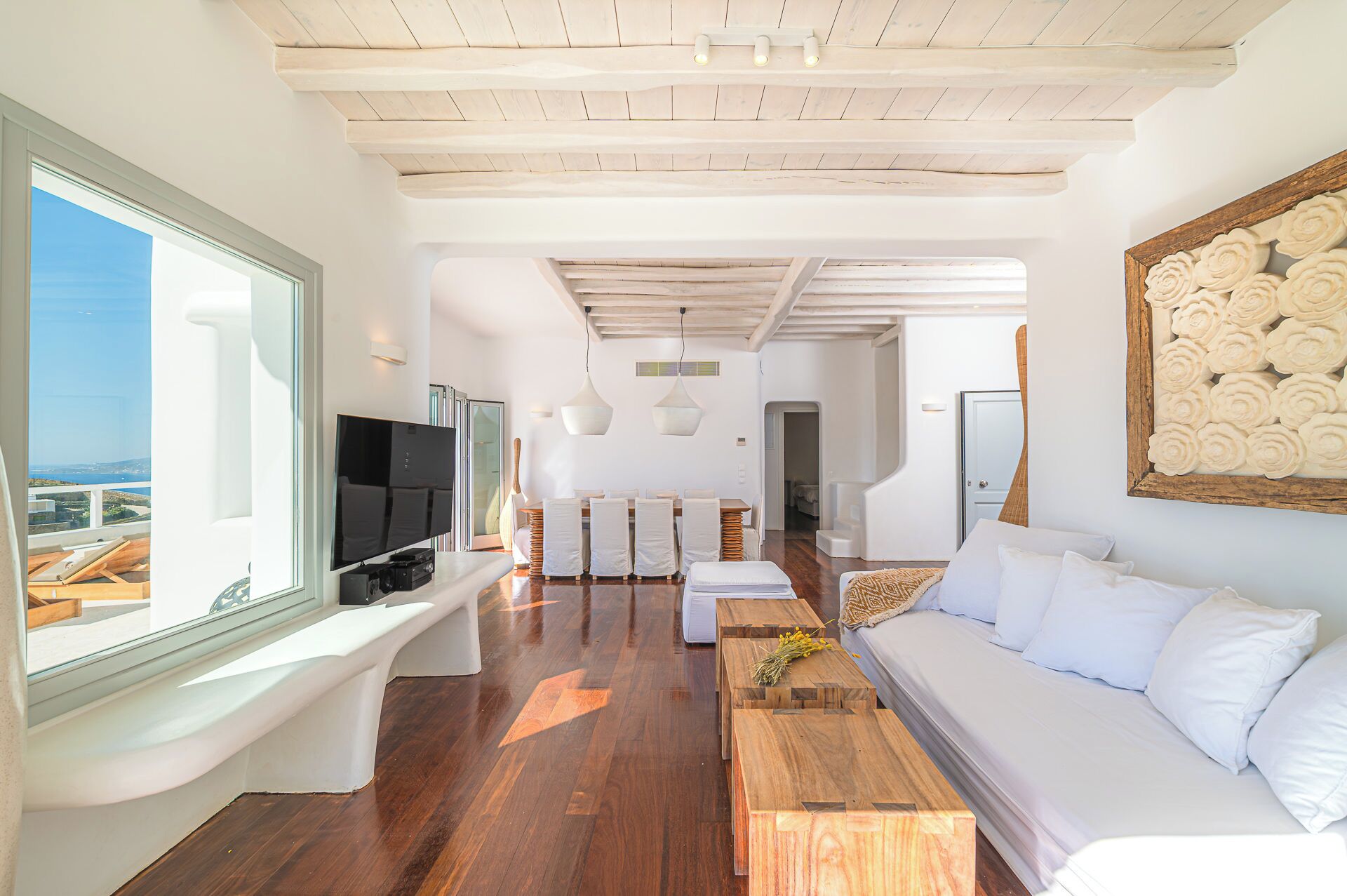 Una sala de estar moderna con un televisor grande, suelo de madera, muebles blancos y vista al mar.