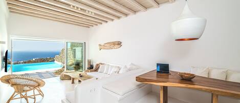 A modern living room with a white sofa, a wooden dining table, and a view of the ocean through large windows.