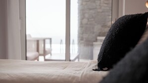A bed with a white sheet and a black pillow, a window with a view of a balcony, and a stone wall in the background. - Anthis Villas Mykonos (Mykonos)