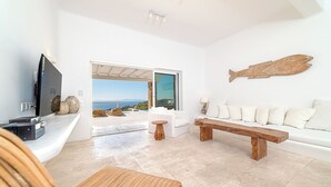 A modern living room with a large TV, a wooden coffee table, a white sofa, and a view of the sea through a glass door. - Anthis Villas Mykonos (Mykonos)