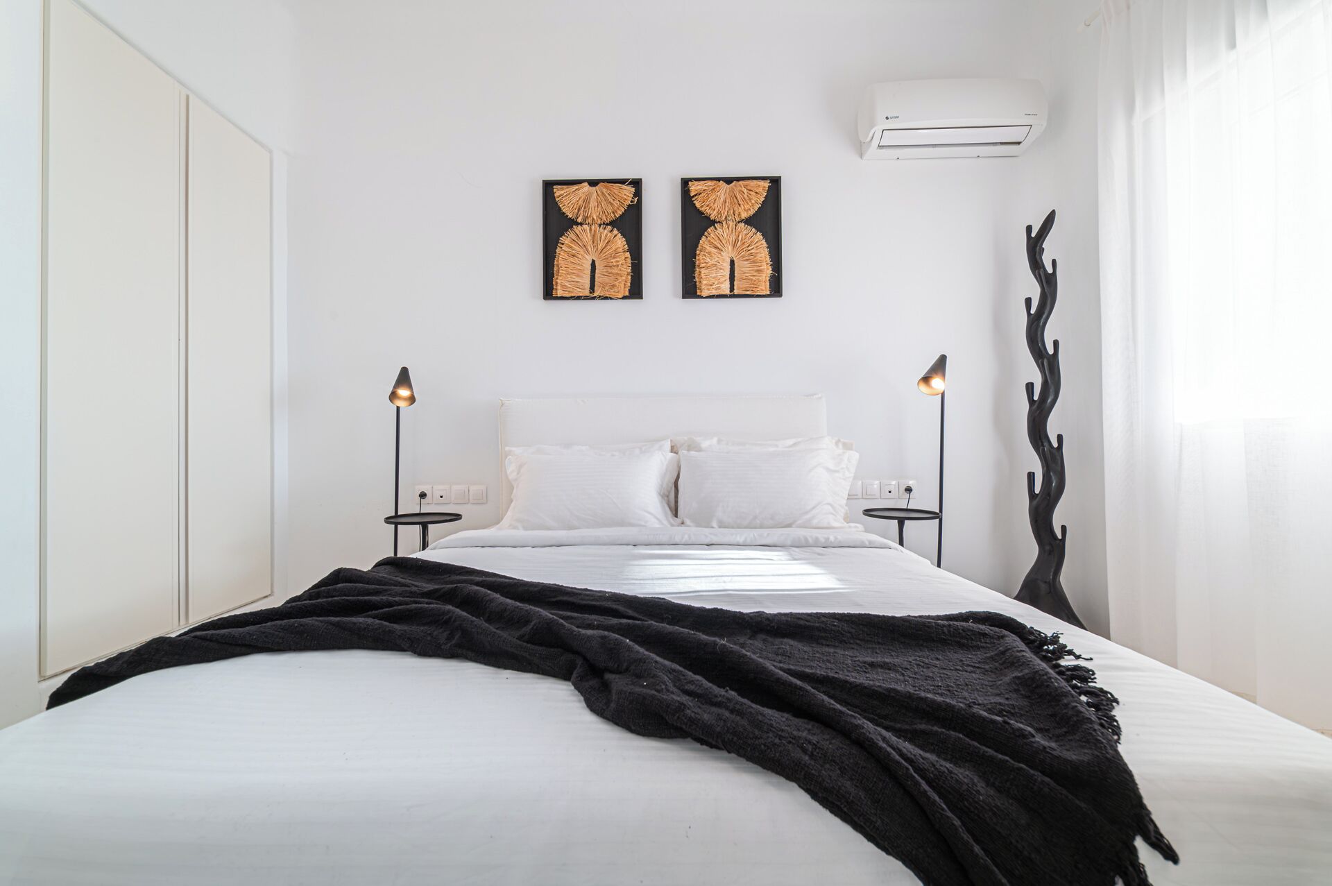 A minimalist bedroom with a large bed, two wall-mounted air conditioning units, and two abstract wall art pieces.