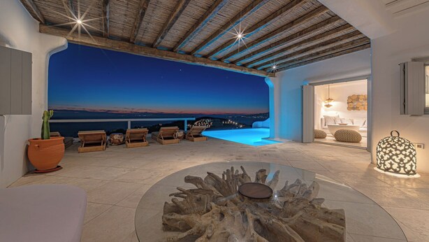 A modern interior with a wooden ceiling, a glass table with a natural wood centerpiece, and a view of the sea at dusk. - Anthis Villas Mykonos (Mykonos)