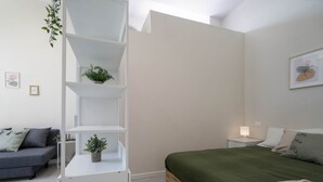 Une chambre à coucher comprenant un lit, une table de chevet, une étagère blanche, un canapé gris et une petite plante en pot.