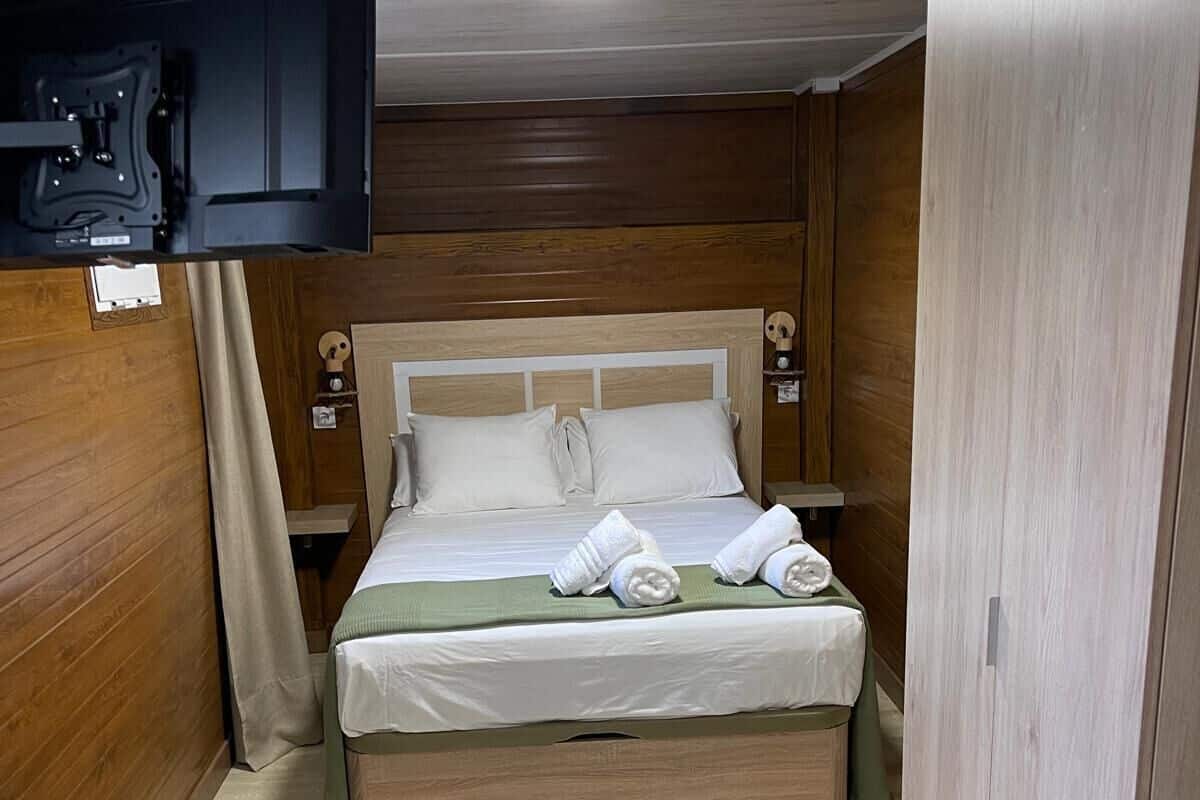 Een netjes opgemaakt bed met kussens en opgerolde handdoeken, een televisie aan de muur bevestigd en een houten garderobe.