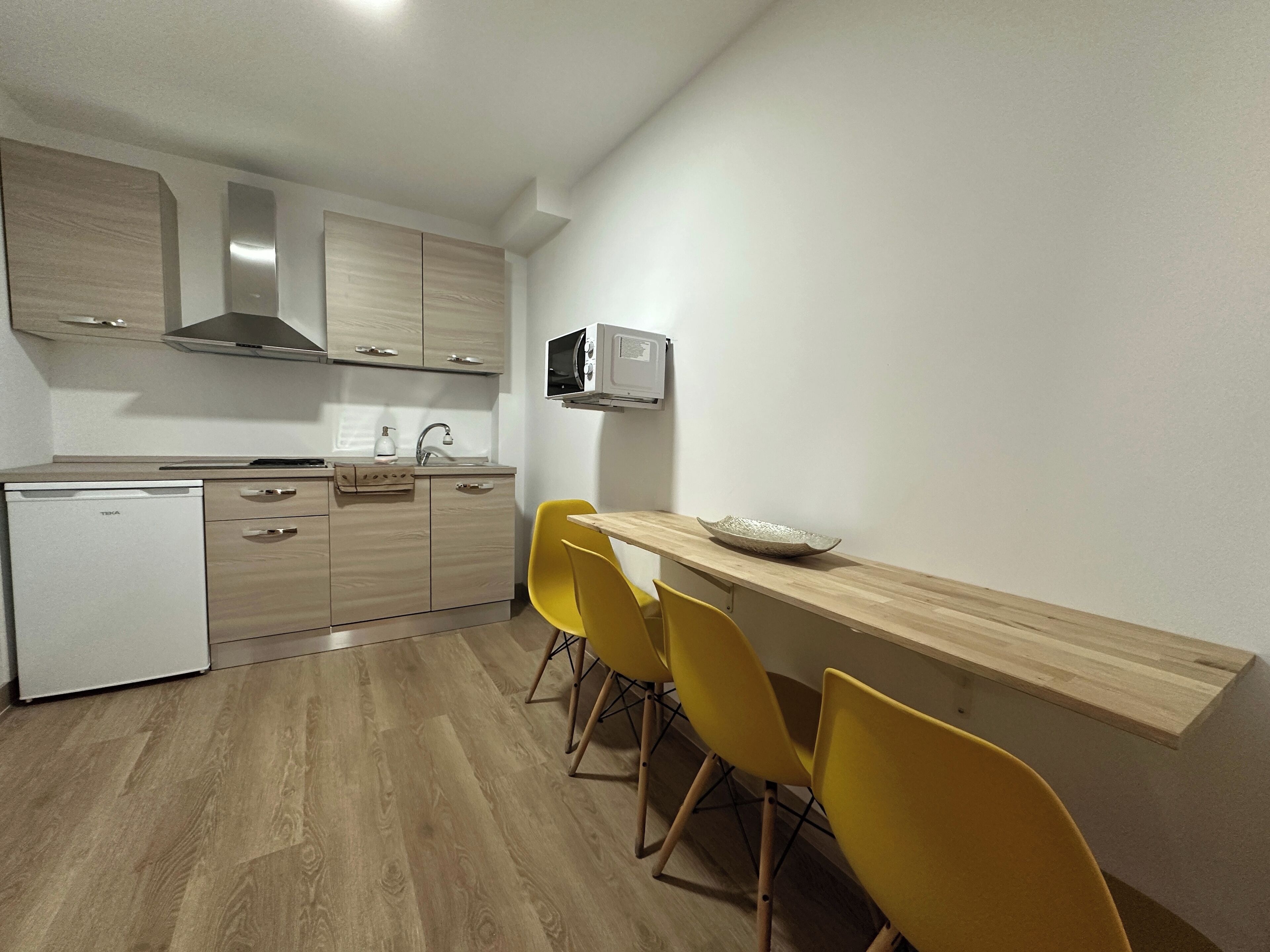 A compact kitchen with wooden cabinets, a built-in microwave, and a sink. There are two yellow chairs and a wooden table.