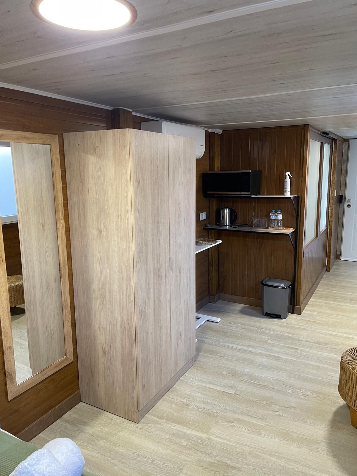 Una cocina compacta con armarios de madera, un microondas, un estante con botellas y un cubo de basura.