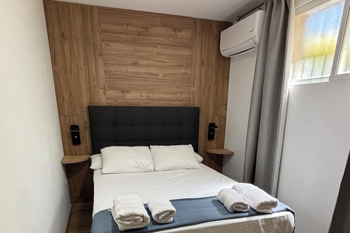 Een netjes opgemaakt bed met witte lakens en handdoeken, een donkere hoofdbord, houten lambrisering aan de muur, een klein nachtkastje en een raam met gordijnen.
