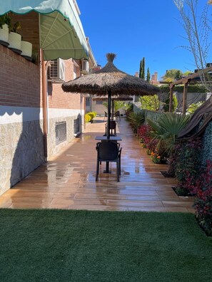 A patio with a table and chairs under a thatched umbrella, surrounded by a garden with various plants and a brick wall. - Habitacion Monteazul (El Casar)