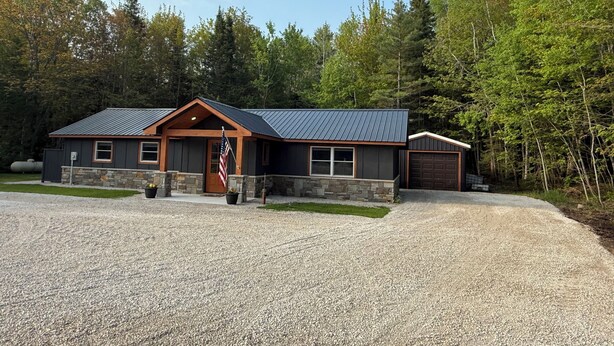 Ein modernes, zweistöckiges Gebäude mit Steinunterbau und Metalldach, umgeben von einer Schotterauffahrt und dichtem Wald.