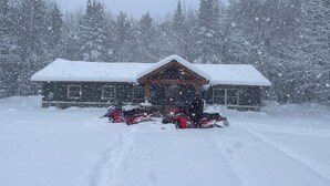 Eine Holzhütte mit verschneitem Dach und einer Holzwand, umgeben von Bäumen und einer schneebedeckten Fläche.