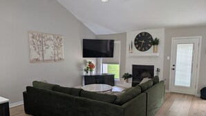 A modern living room with a sectional sofa, a flat-screen TV, a fireplace, and a large clock on the wall. - Beautiful 3/2 Home, quiet location, close proximity to Texas A&M University (Bryan)
