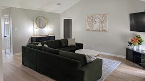 A modern living room with a sectional sofa, a small round table, and a flat-screen TV mounted on the wall. - Beautiful 3/2 Home, quiet location, close proximity to Texas A&M University (Bryan)