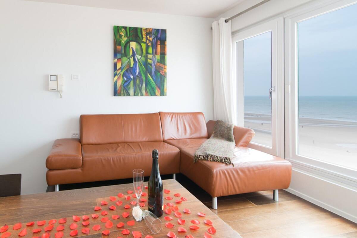Un salon moderne avec un canapé en cuir marron, une table en verre ornée de pétales de rose, une bouteille et un verre. Un tableau abstrait aux couleurs vives est accroché au mur, et une grande fenêtre offre une vue sur la plage.
