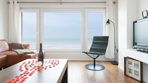 A modern living room with a wooden floor, a leather sofa, a green chair, a dining table with rose petals, and a TV. - Studio in Ostend Near Beach and Promenade (Ostend)