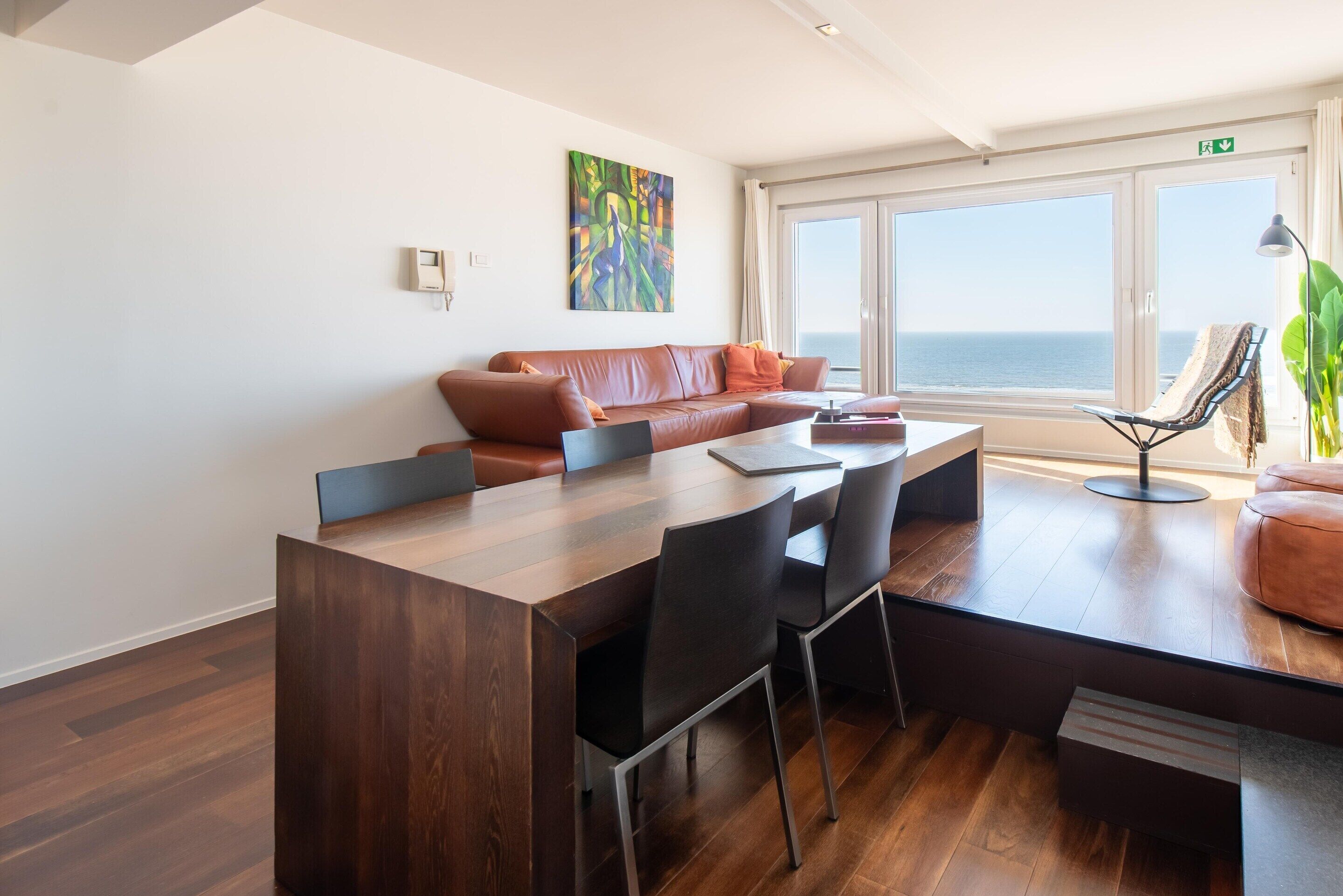 Una sala de estar moderna con una mesa de centro de madera, sillas negras, un sofá de cuero marrón y un amplio ventanal con vistas al mar.