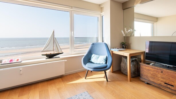 Un salon moderne avec une grande fenêtre donnant sur la mer, un fauteuil bleu, un bureau en bois et un téléviseur.