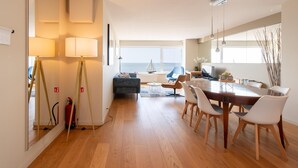 Un salon moderne avec un plancher en bois, un coin repas avec une table en bois et des chaises blanches, un canapé bleu et une vue sur la mer par de grandes fenêtres.