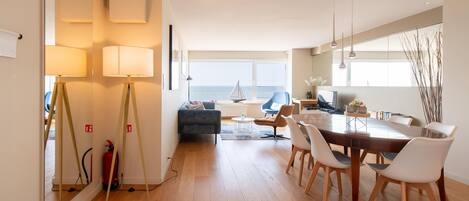 A modern living room with a wooden floor, a dining area with a wooden table and white chairs, a blue sofa, and a view of the sea through large windows.