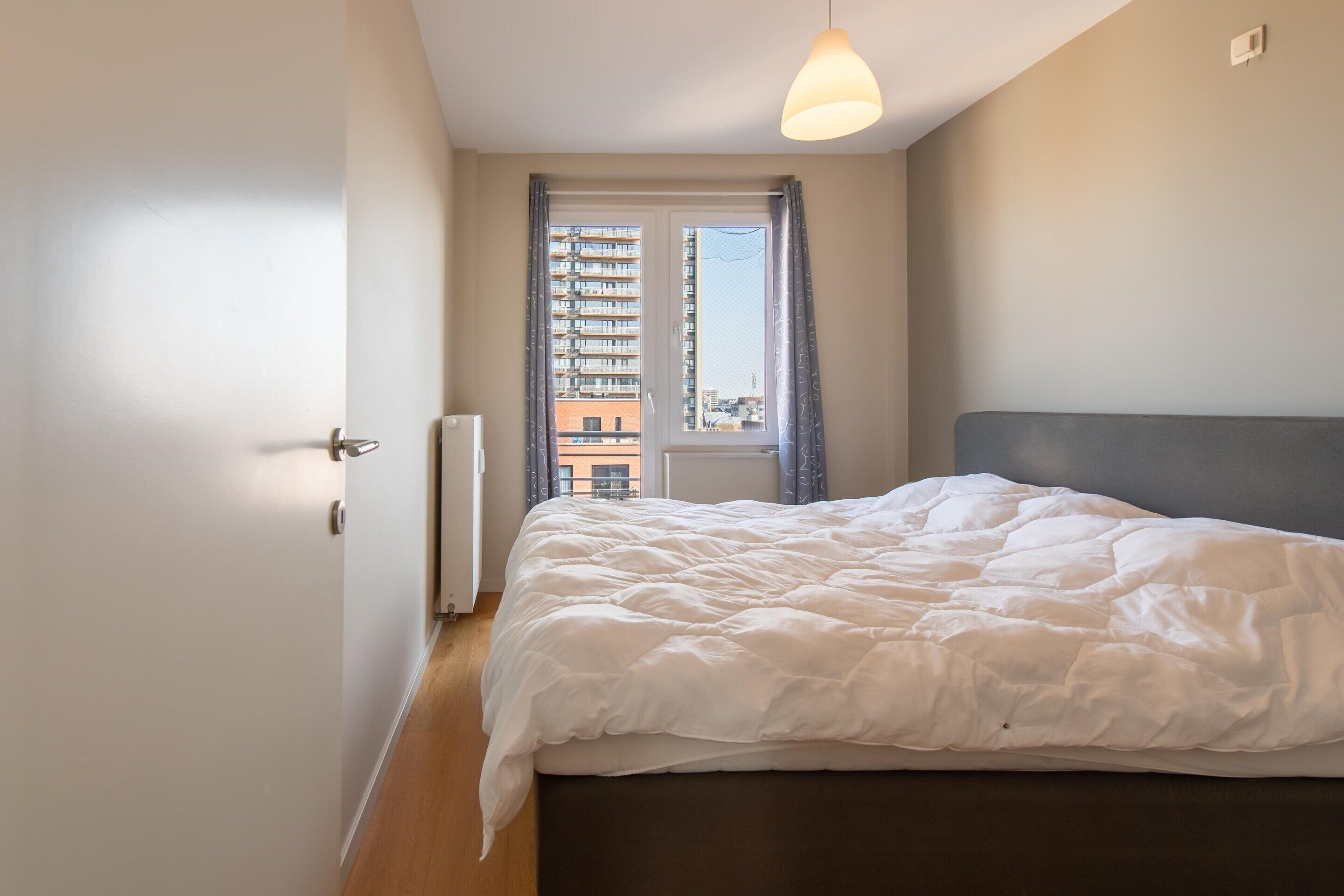 A bedroom with a bed, a window with curtains, a radiator, and a view of buildings outside.