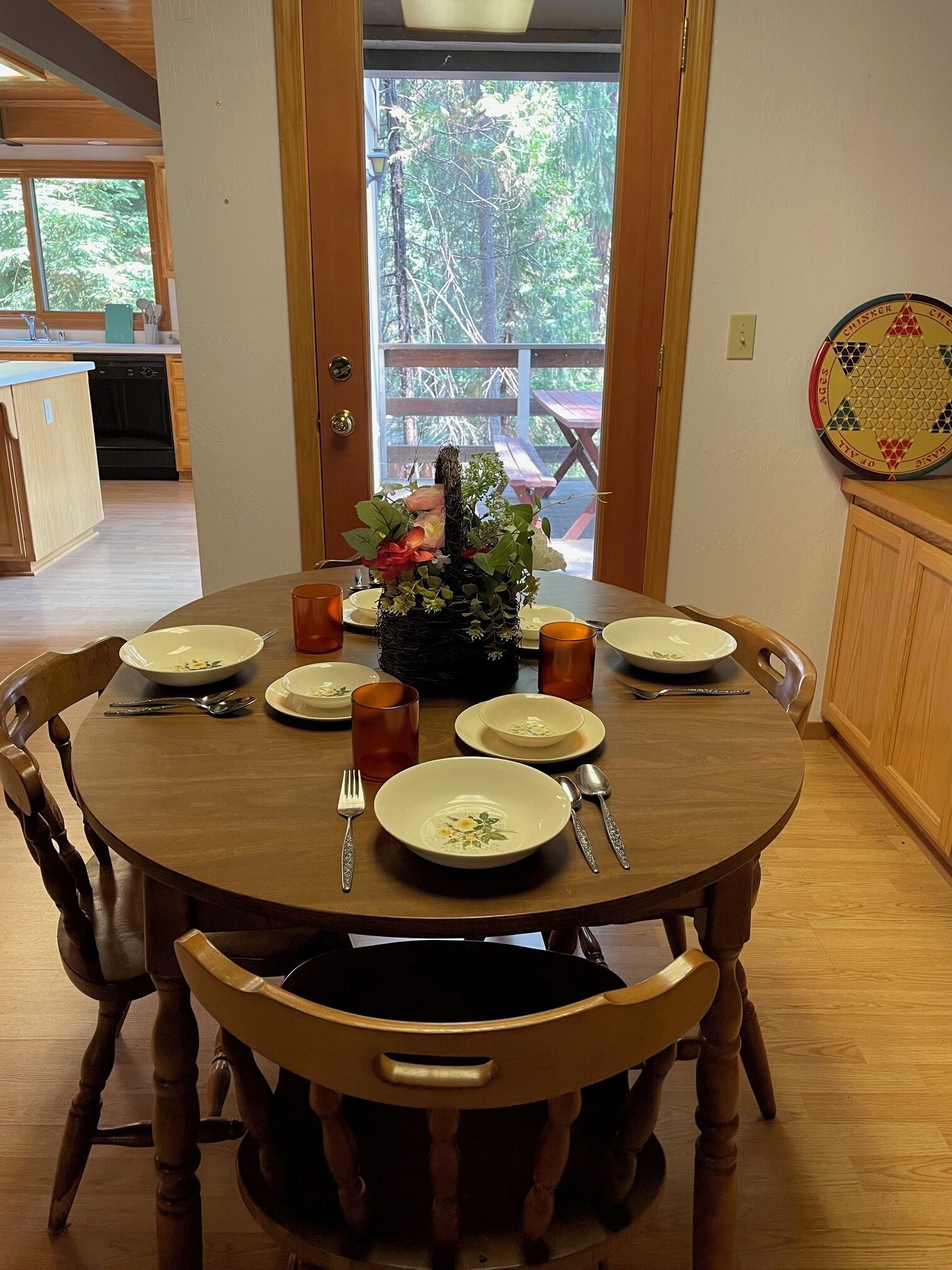 A dining table set for six with a centerpiece, surrounded by wooden chairs. A wooden cabinet and a door leading to an outdoor area are visible.