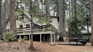 A two-story cabin with a covered porch and a wooden deck, surrounded by tall trees in a forested area. - GREAT FAMILY LOCATION!  Peace in the Trees N Mace (Pioneer)