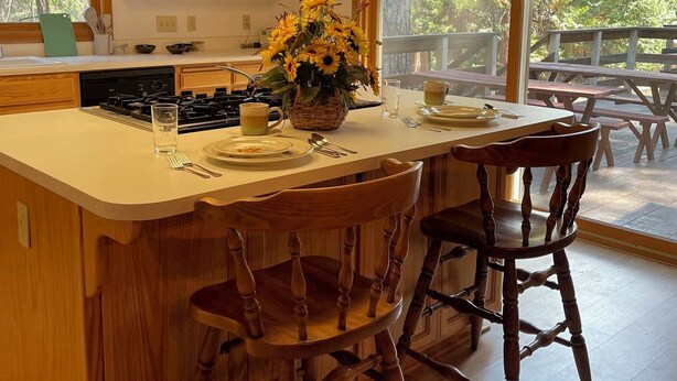 A kitchen with wooden cabinets, a central island with bar stools, a dining table outside, and a large window with a view of trees. - GREAT FAMILY LOCATION!  Peace in the Trees N Mace (Pioneer)