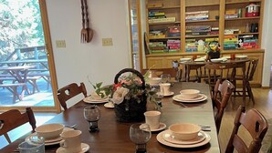 A dining area with a wooden table set for four, surrounded by chairs. A basket of flowers is placed on the table, and there are various board games and books on shelves in the background. - GREAT FAMILY LOCATION!  Peace in the Trees N Mace (Pioneer)