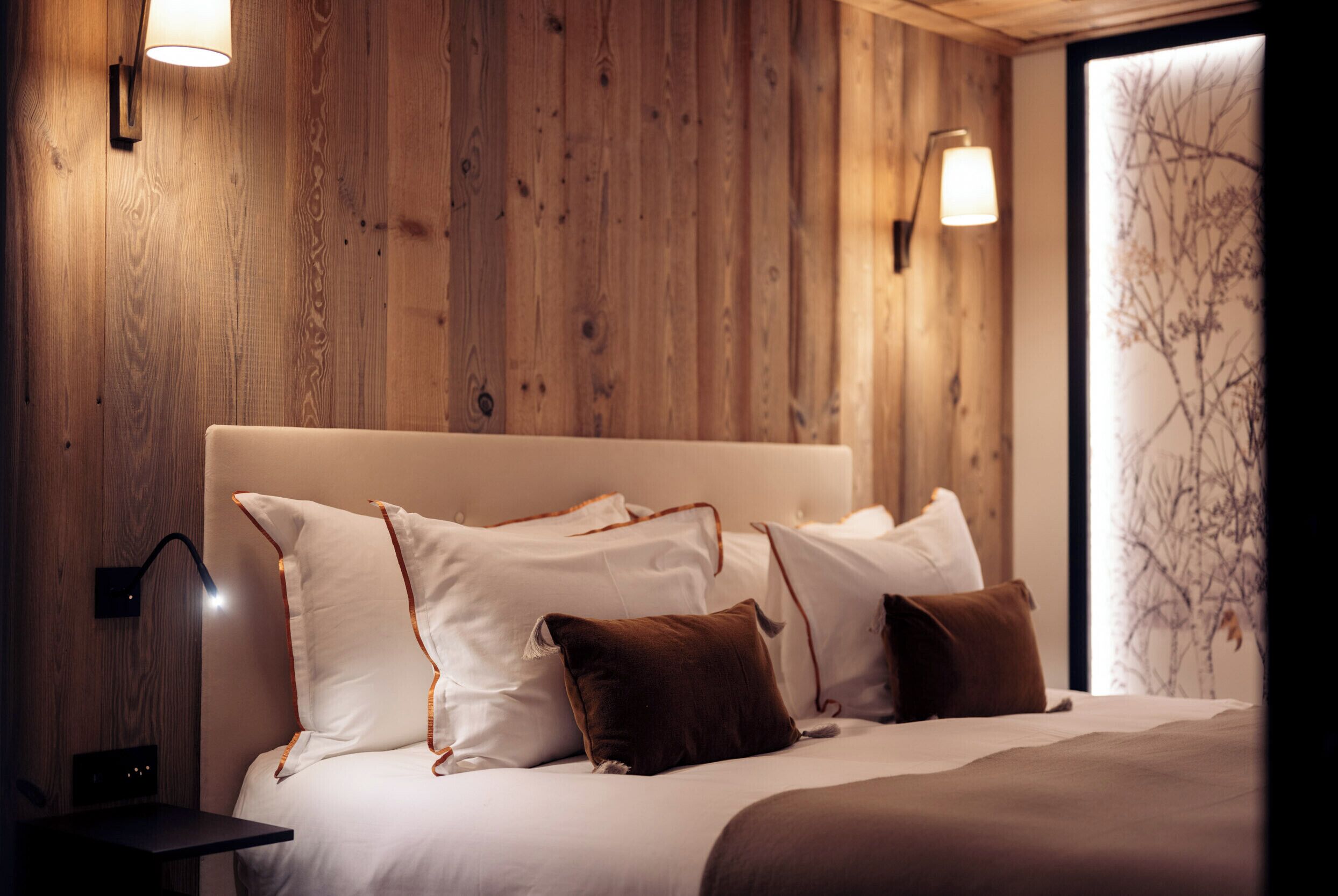 A bedroom with a wooden wall, a bed with white and brown bedding, and two wall-mounted lamps.