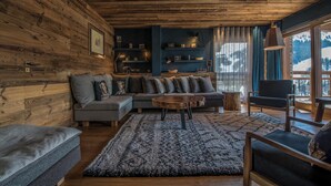 Una sala de estar acogedora con techo de madera, un sofá modular, una mesita de centro redonda y vistas a montañas a través de amplios ventanales.