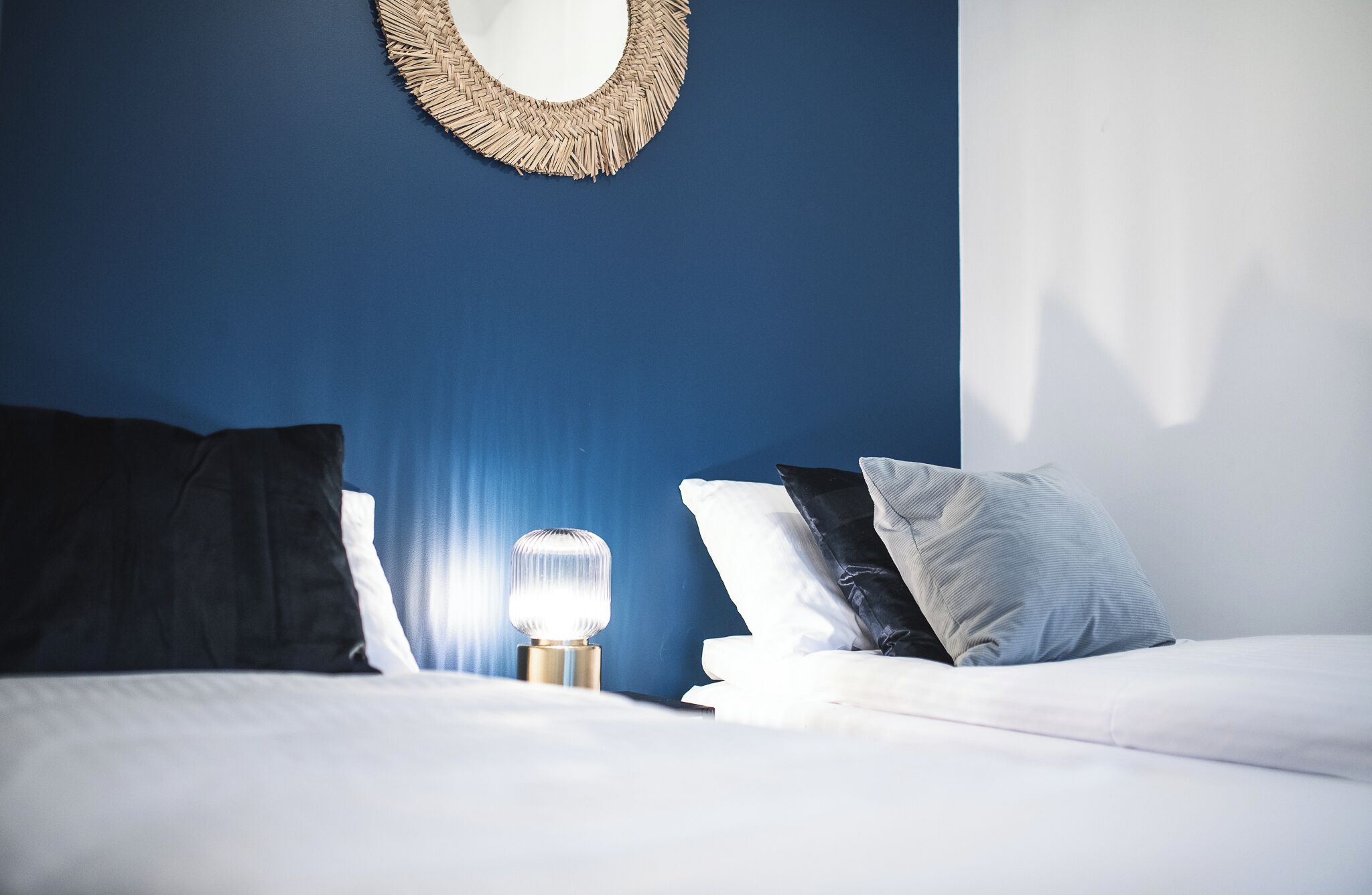 A neatly made bed with white linens, a bedside lamp, and a woven wall hanging.