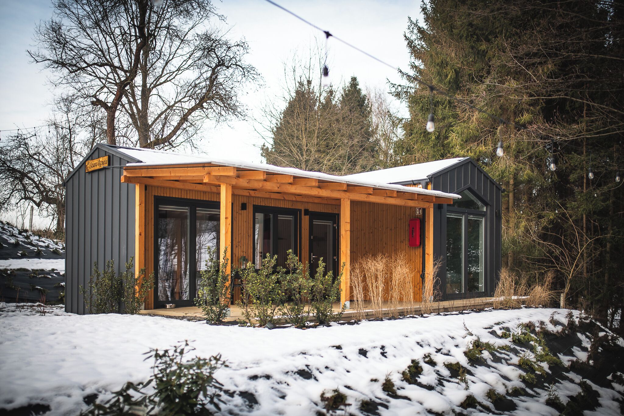 A modern, two-story cabin with a wooden porch and large windows, surrounded by snow and trees.