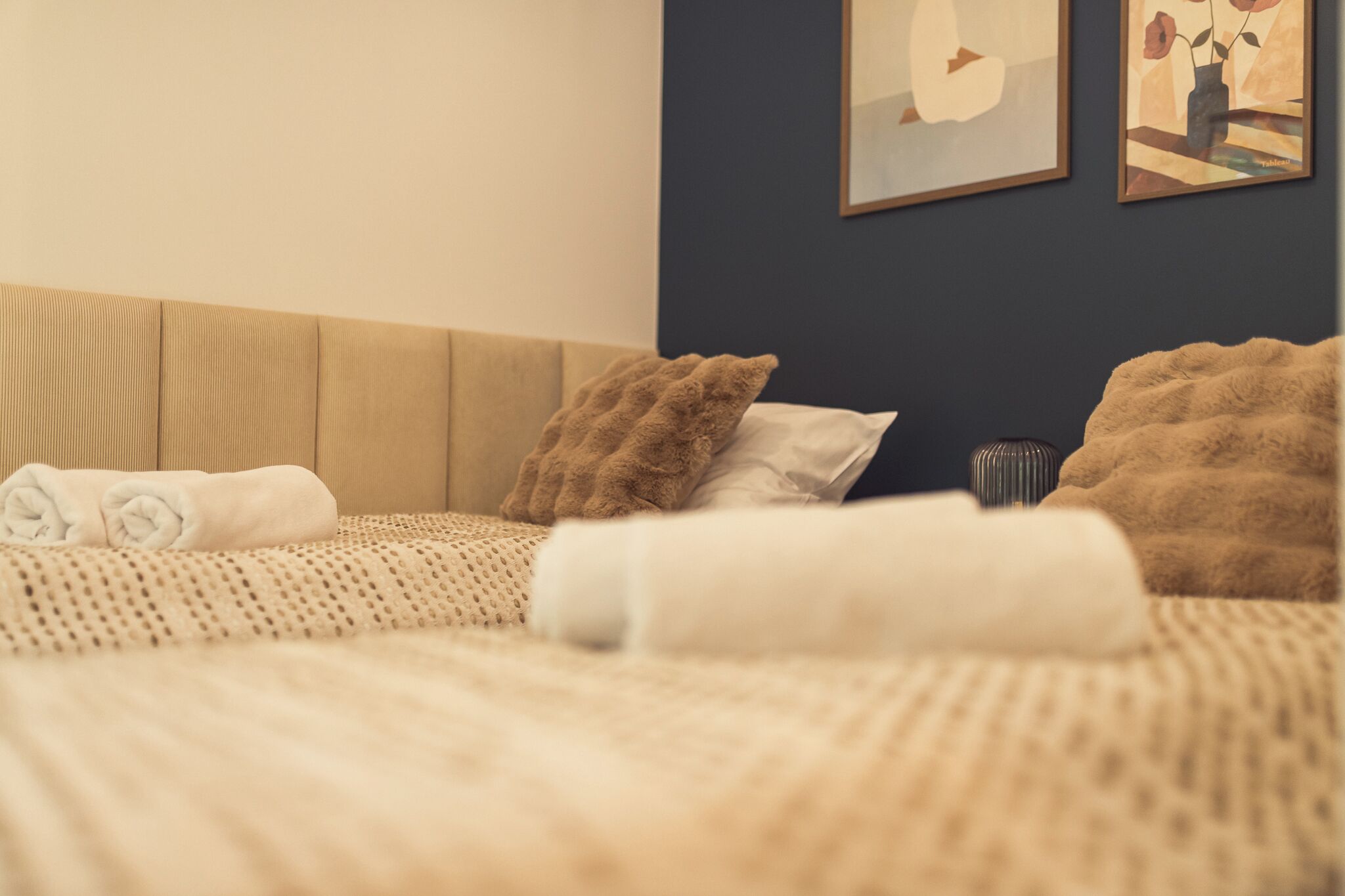 Una cama bien hecha con toallas enrolladas, una manta texturizada y una pared oscura con dos obras de arte enmarcadas.