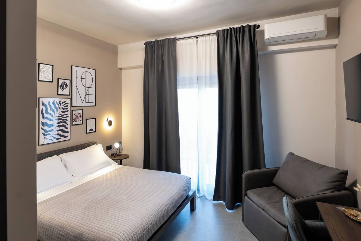 A hotel room with a bed, a sofa, a wall-mounted air conditioner, and framed artwork on the wall.