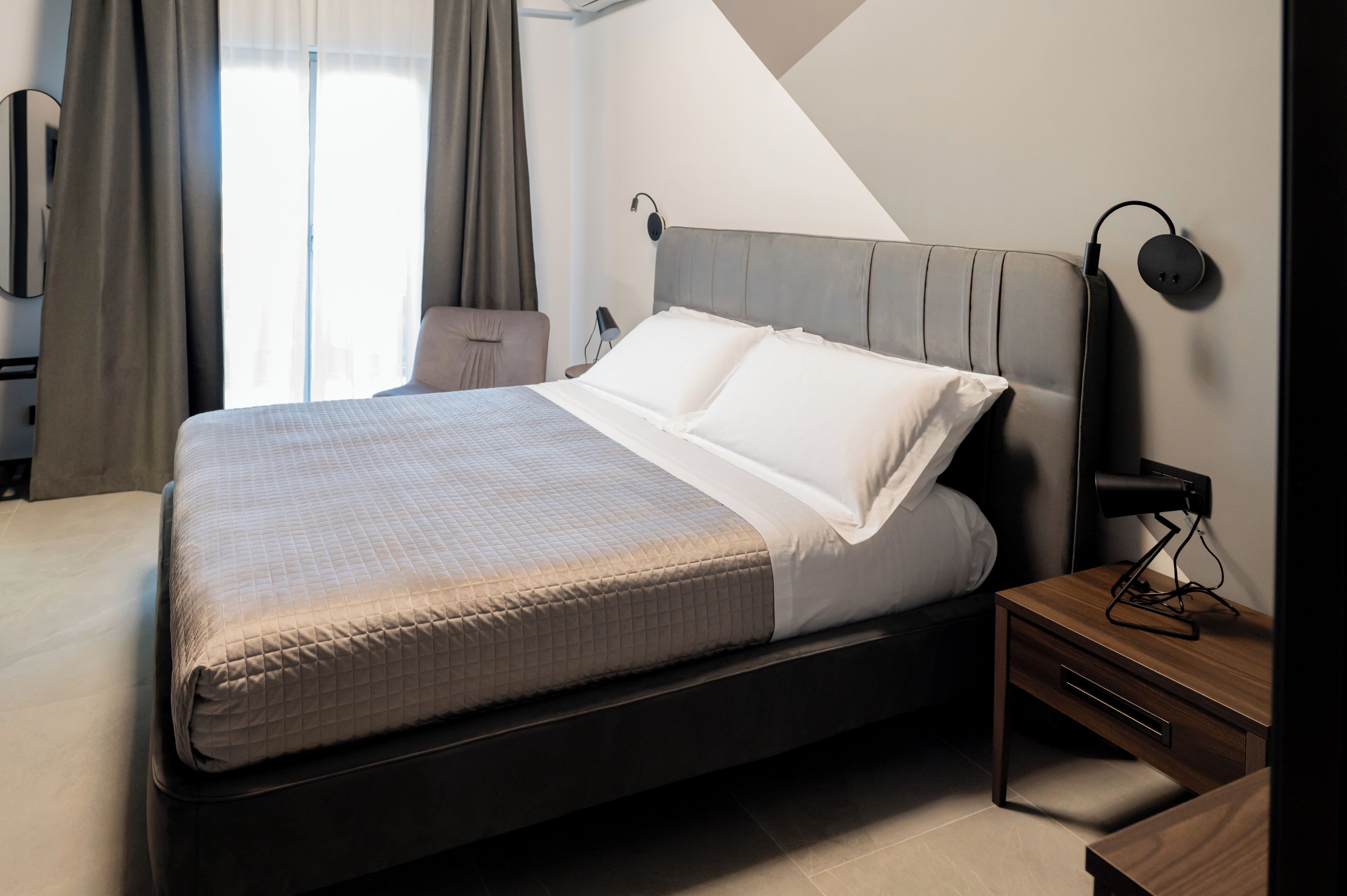 A modern hotel room with a large bed, a bedside table, a lamp, and a view of the city through the window.