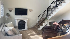 A living room with a fireplace, a sofa, a brown leather chair, a wooden coffee table, and a staircase with a black railing.