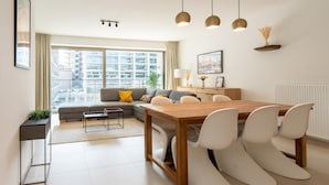 Un salon moderne avec une table à manger en bois, des chaises blanches, un canapé gris et une grande fenêtre donnant sur la ville.