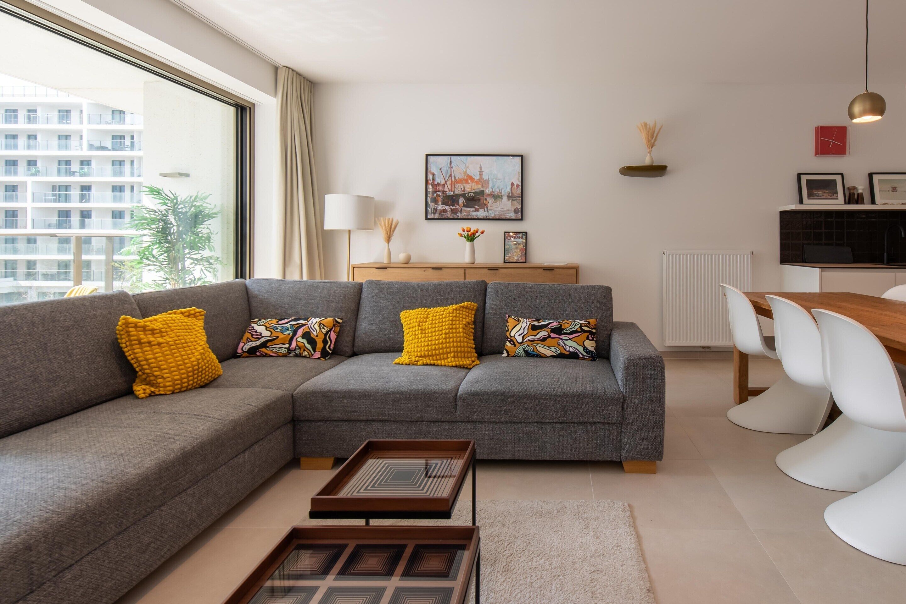 A modern living room with a grey sectional sofa, a wooden coffee table, and a dining area with white chairs.