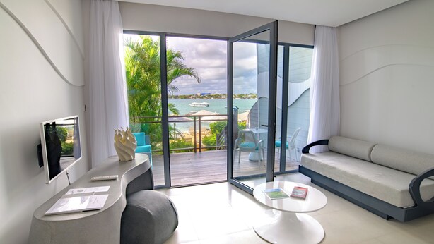Un salon moderne avec un canapé, une table basse et un téléviseur, donnant sur une terrasse offrant une vue sur la mer et les palmiers.