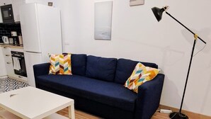 Ein kompakter Wohnbereich mit einem weißen Sofa, einem kleinen weißen Tisch, einer schwarzen Stehlampe und einer Küchenzeile mit Mikrowelle und Kühlschrank.